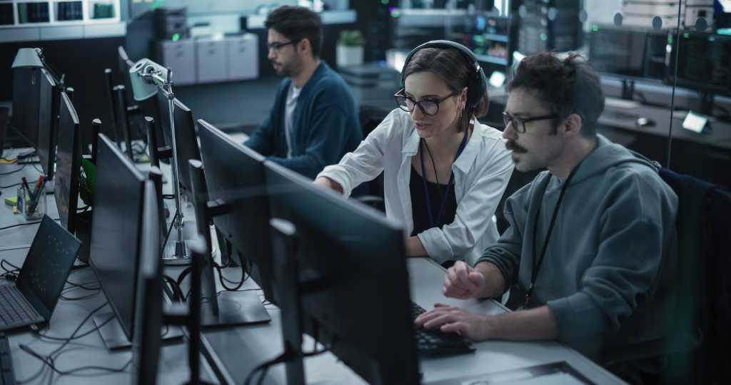 3807978-0-93132300-1739996157-shutterstock_2287185555-1 Young Team of Specialists Working on Desktop Computers and Having a Conversation at a Workplace. Female and Male Software Developers Discussing a Solution for Their Artificial Intelligence Project