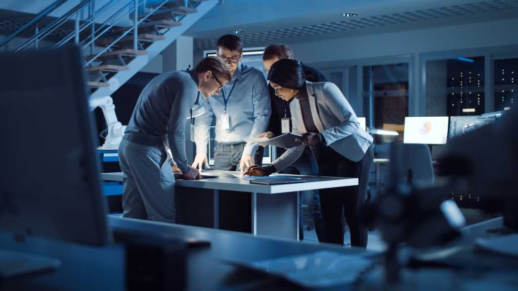3812589-0-73601800-1740387761-shutterstock_1699780492-1 Diverse Team of Electronics Development Engineers Standing at the Desk Working with Documents, Solving Project Problems Late at Night. Specialists Working on Ultra Modern Industrial Design.