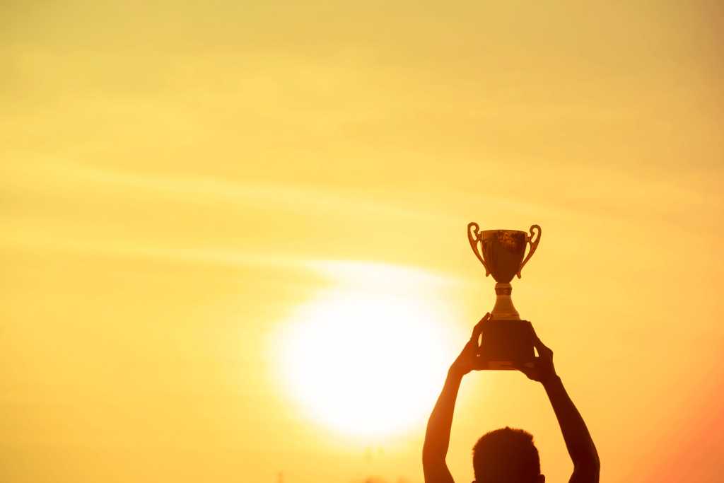 3814855-0-28829100-1738777031-shutterstock_1024091266-100963204-orig-1 Winner holding golden trophy cup above head sun and golden sky in background