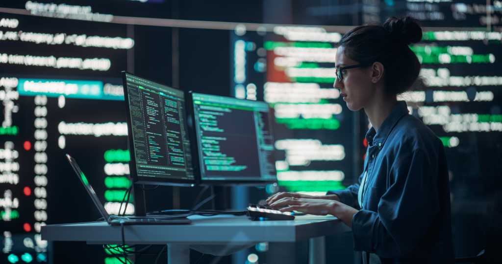 3839444-0-98017200-1741251708-shutterstock_2324952341-1 Female Developer Thinking and Typing on Computer, Surrounded by Big Screens Showing Coding Language. Professional Programmer Working in an Office, Running Coding Tests. Futuristic Programming
