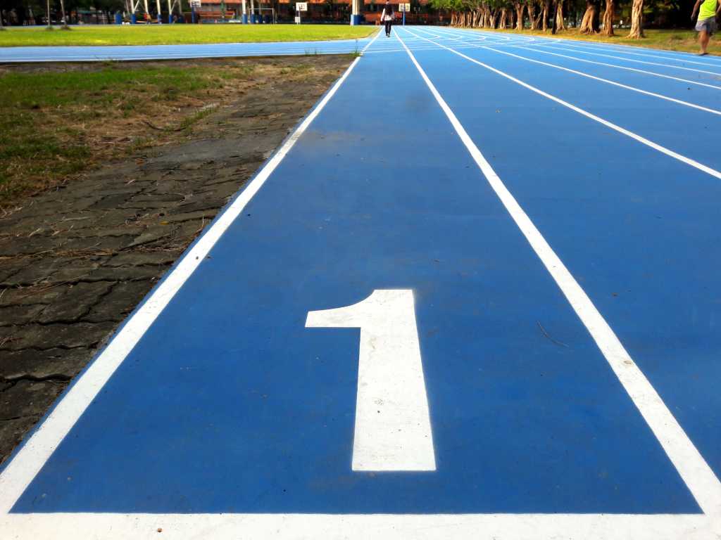 3844448-0-37345600-1741856498-shutterstock_2307918973-1 Number 1 / number one lane of running track, white number and white lane lines on blue surface. Low level view with grass runners trees visible in the distance.