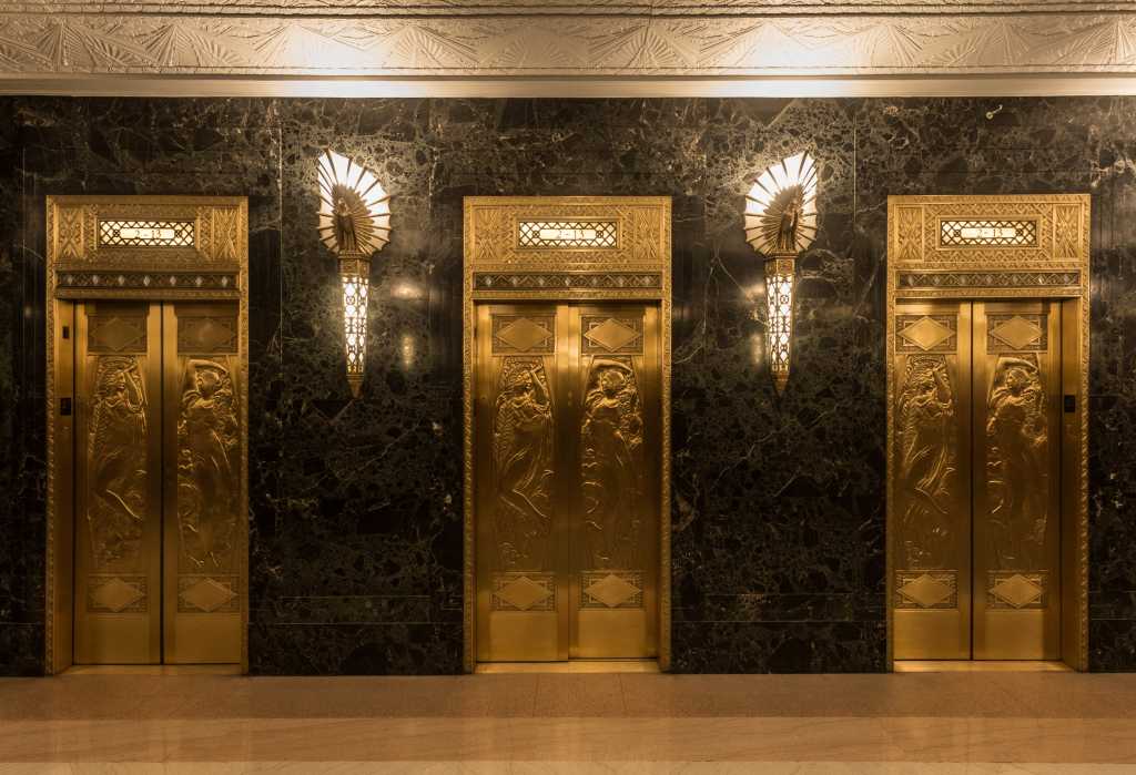 3847079-0-39342900-1742461286-shutterstock_734738725-1 Dramatic golden elevators in fancy urban skyscraper building with relief sculptures and marble wall with lights and sconces