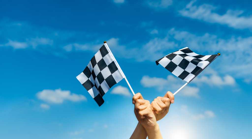 3849654-0-48898100-1742461276-shutterstock_2358602889-1 Two checkered flags against a blue sky, held by crossed hands