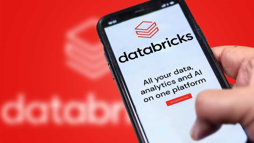 3842059-0-36812700-1741692882-databricksphone-1 A photograph of a Databricks website on a phone being held in front of a larger, blurry version of the Databricks logo.