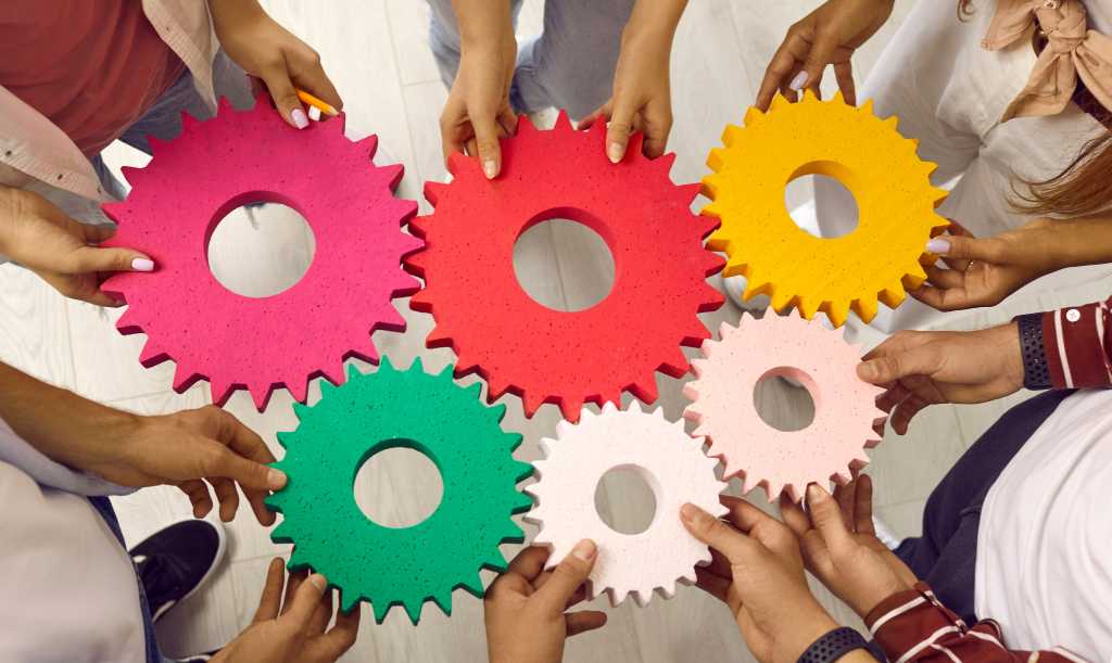 3961824-0-92074800-1744707674-shutterstock_2244247863-1 Team or group of men and women standing in circle joining gears holding colorful cogs. Cropped shot. Teamwork, integration, business, education, success concept background