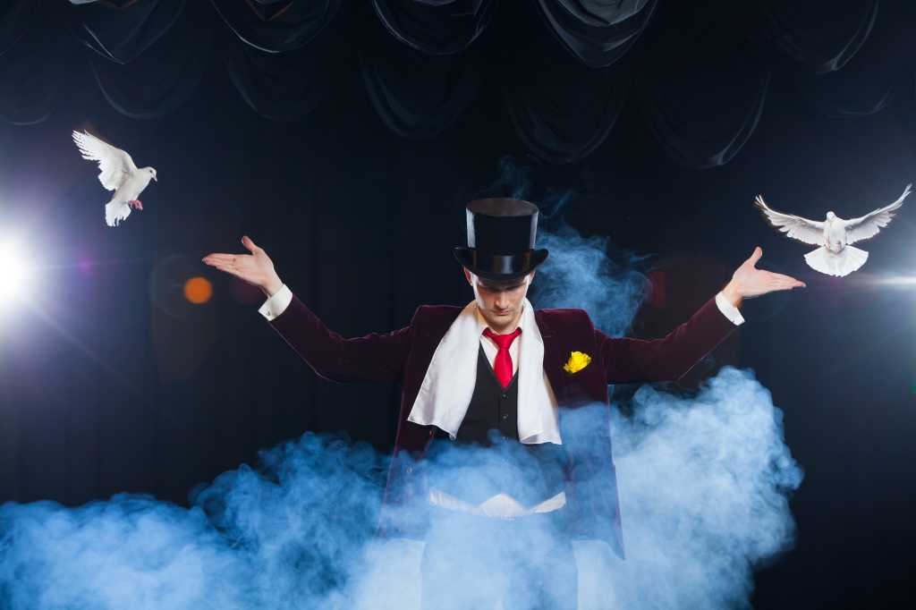 3984227-0-19347000-1747213321-shutterstock_762306028-1 Magician with two flying white Doves on a black background shrouded in mysterious blue smoke